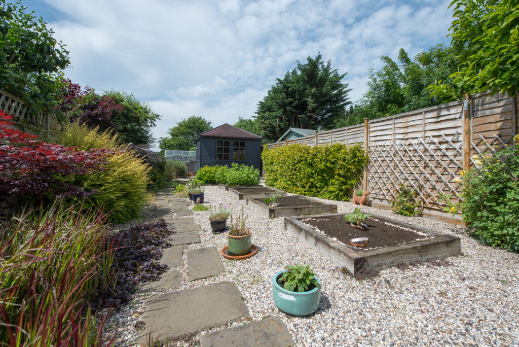 A view looking down a back garden of a home with paving slaps and gravel, pea shingle, wooden railway sleeper flower bed, vegetable patch, potted plants, timber fence and grey summerhouse, shed with tiled roof on a warm sunny day