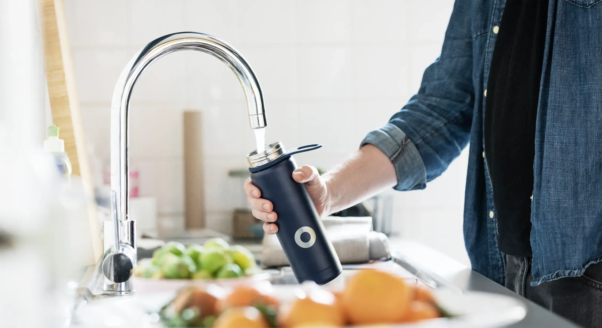 Torso and forearm filling metal water bottle with purified water from kitchen faucet.